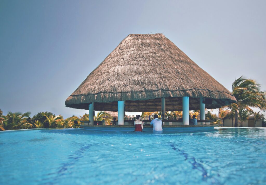 Casal sentado em cadeiras de bar dentro de uma piscina, em frente a um bar molhado com teto de palha e colunas brancas, em um dia de sol.