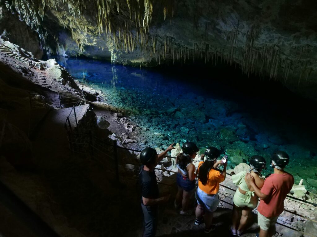Turistas com capacetes em uma plataforma de observação dentro da Gruta do Lago Azul em Bonito, admirando o lago de águas azuis e cristalinas com estalactites no teto da caverna.