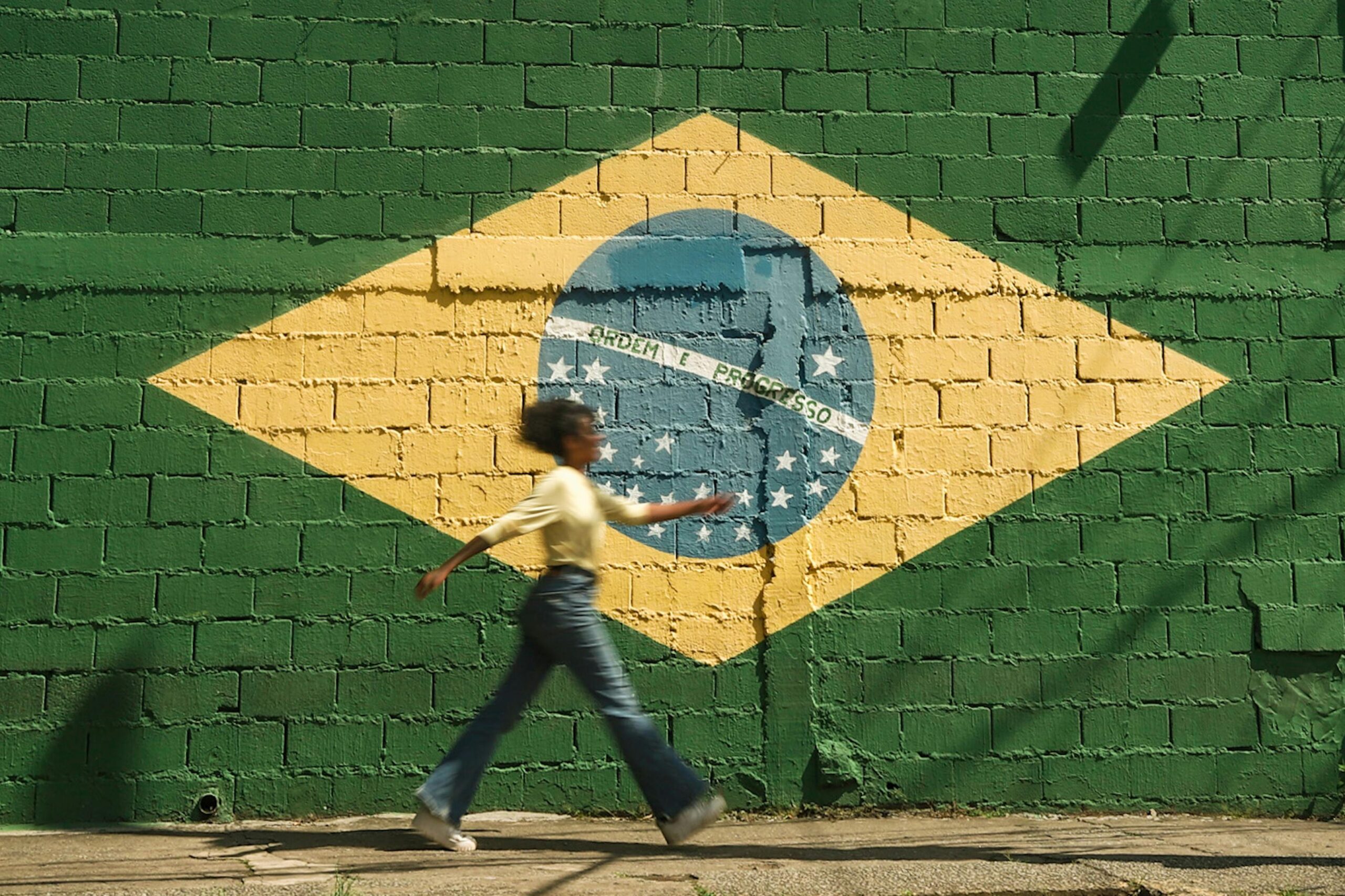 Uma pessoa caminha em frente a um muro de tijolos pintado com a bandeira do Brasil, representando a jornada e as diversas experiências de viagem pelo país.