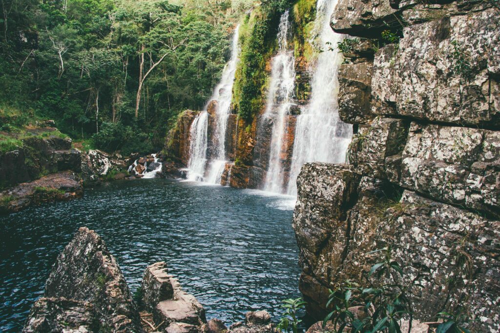 Uma cachoeira de múltiplas quedas d'água desce por rochas escuras e musgosas, formando um poço de águas escuras cercado por vegetação densa e rochas em primeiro plano, na Chapada dos Veadeiros.
