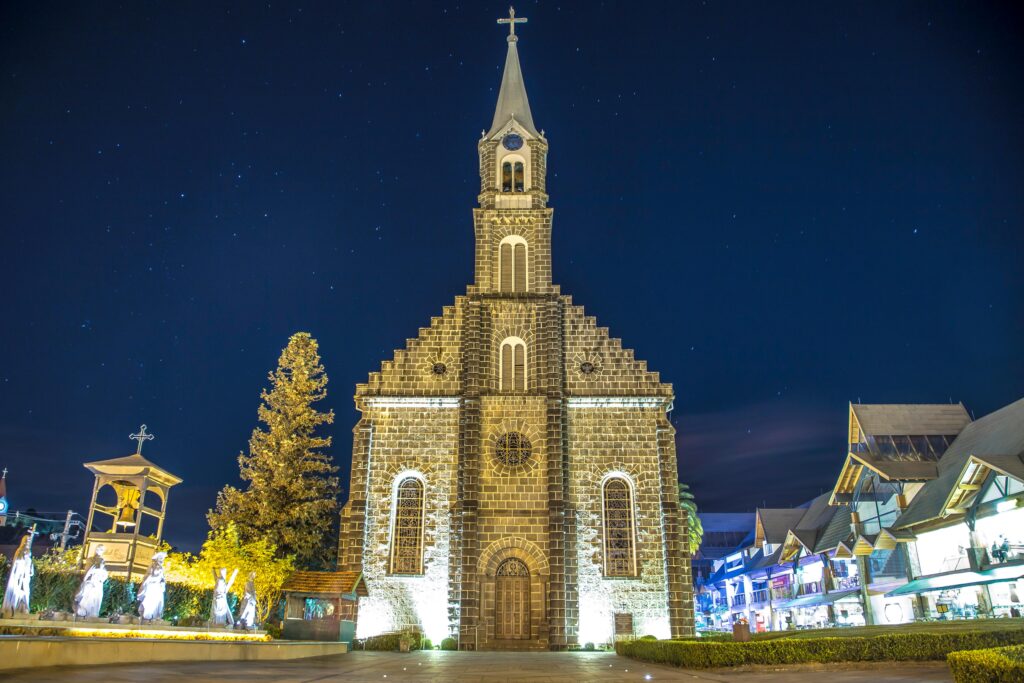 Vista noturna da Igreja Matriz São Pedro, em Gramado, com sua fachada de pedra e torre iluminadas, contrastando com o céu escuro e estrelado.