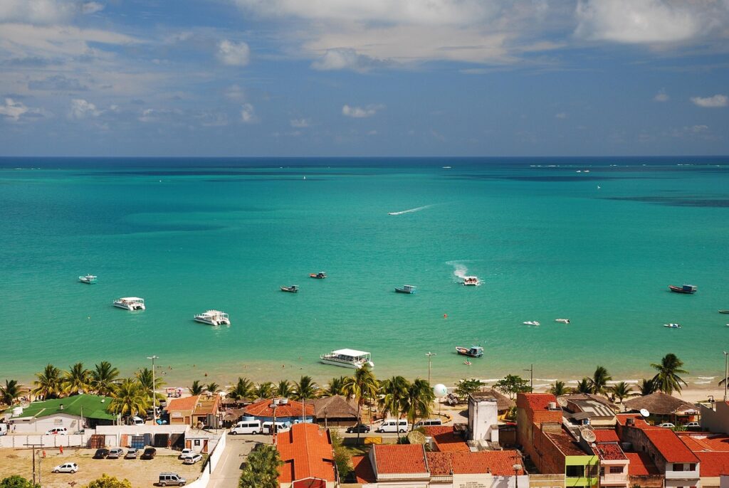 Vista panorâmica da praia de Maragogi, em Alagoas, com o mar de águas calmas e de cor azul-turquesa, barcos flutuando e a orla com coqueiros e construções à beira-mar.