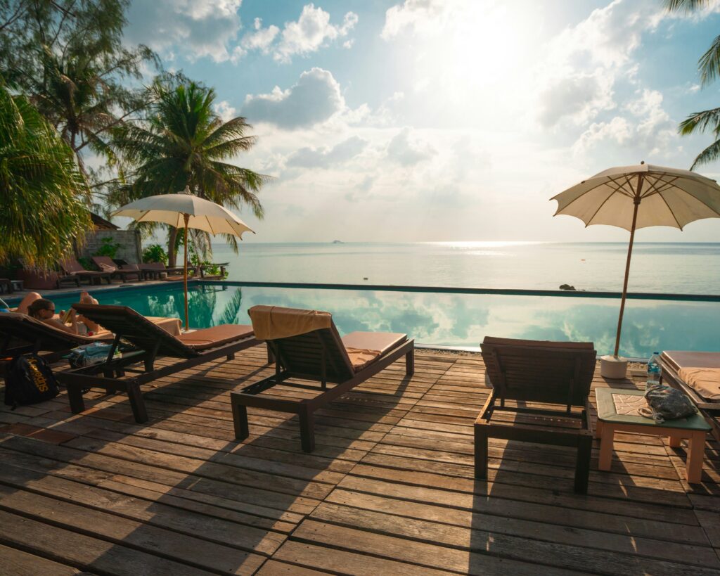 Piscina de borda infinita com vista para o mar, cercada por um deck de madeira com espreguiçadeiras e guarda-sóis, e coqueiros ao fundo.