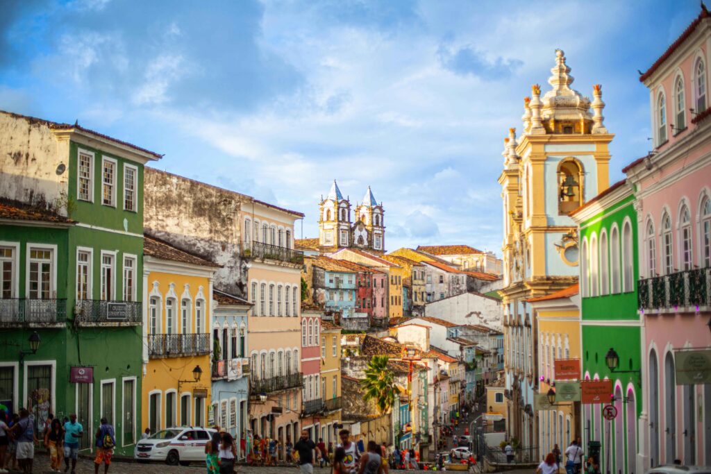 Rua de paralelepípedos no Pelourinho, em Salvador, ladeada por casarões coloniais coloridos e bem preservados, com pessoas caminhando e uma igreja barroca imponente ao fundo sob um céu parcialmente nublado.
