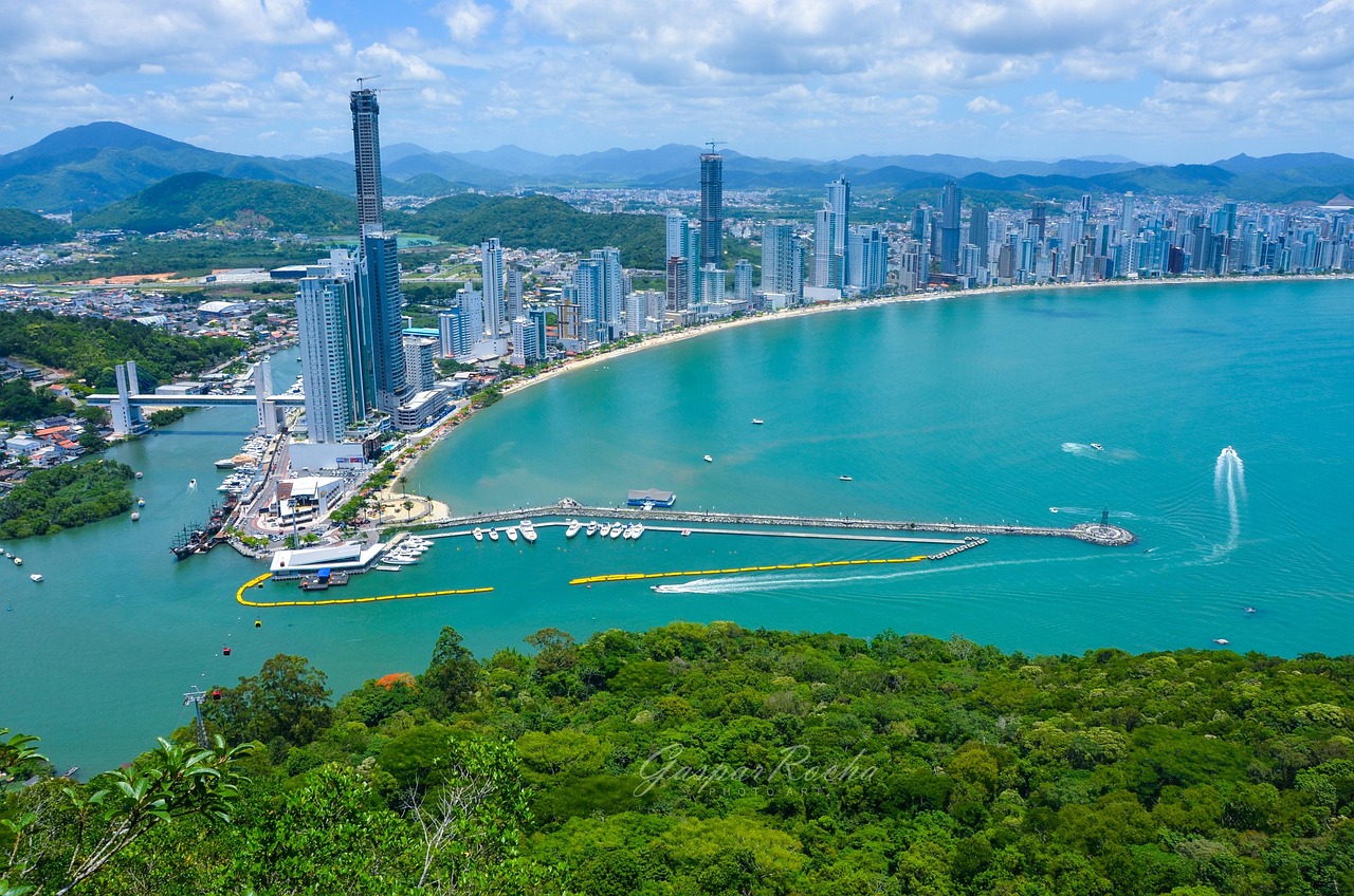 Vista panorâmica aérea de Balneário Camboriú ao entardecer, mostrando a baía da Praia Central, a Ilha das Cabras, diversos arranha-céus na orla e a roda-gigante (FG Big Wheel).