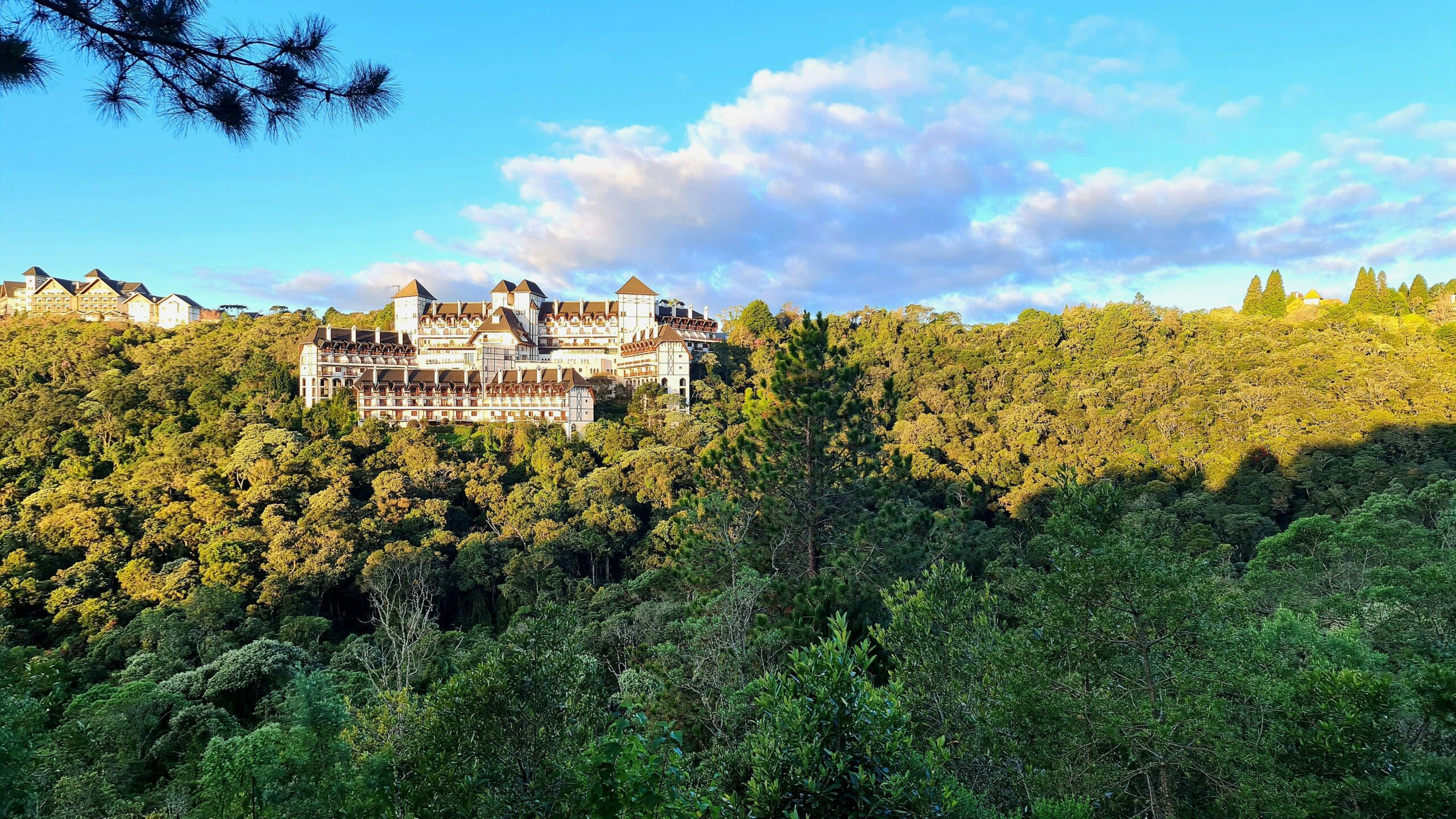 Vista panorâmica do imponente Palácio Boa Vista, em Campos do Jordão, com arquitetura em estilo normando, cercado por uma densa floresta de árvores verdes e sob um céu azul.