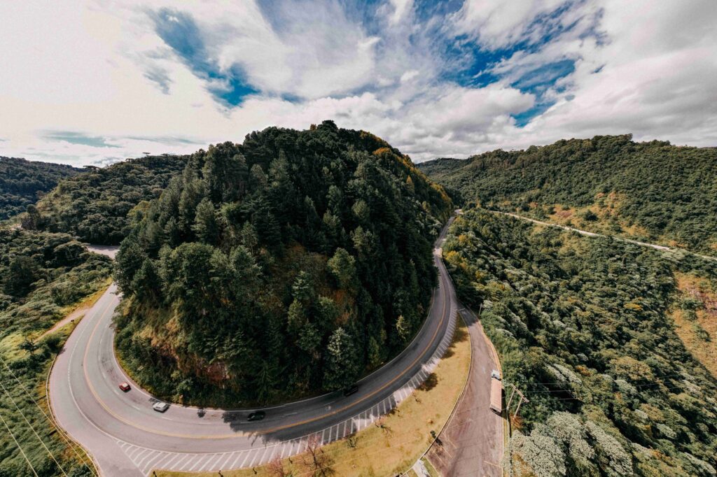 Estrada sinuosa (curvas acentuadas) com asfalto escuro subindo a serra coberta por vegetação verde densa, indicando o caminho para a cidade de Campos do Jordão, em dia de céu com nuvens brancas.