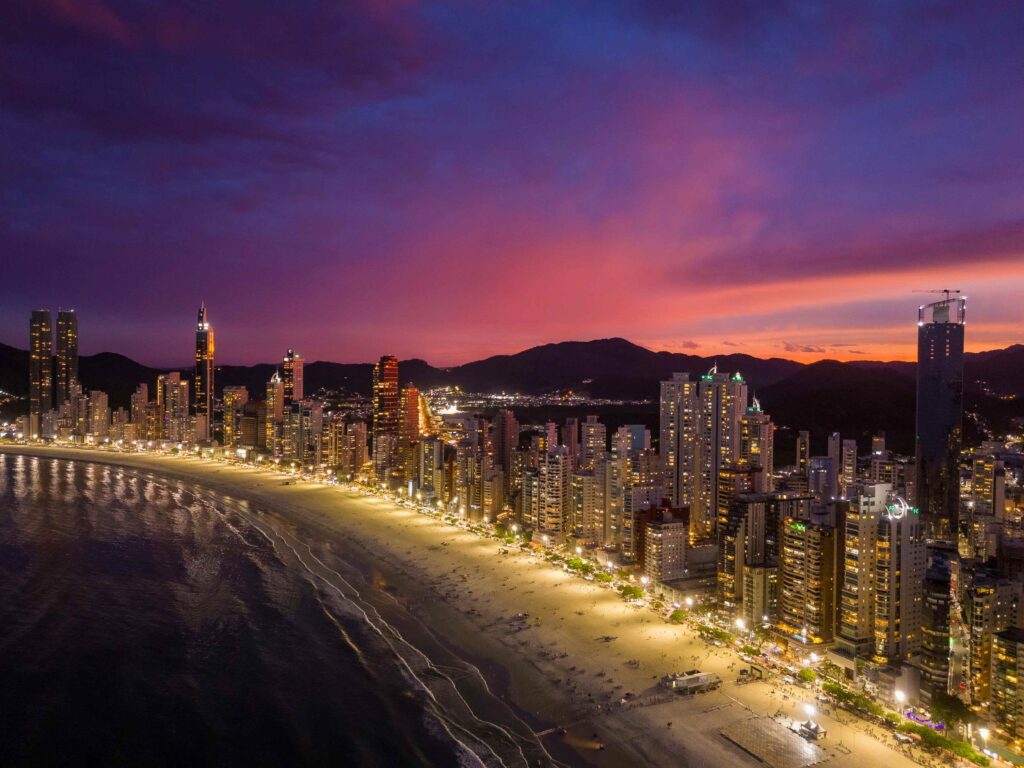 Vista aérea noturna da Praia Central de Balneário Camboriú com um intenso céu roxo e rosa ao pôr do sol, mostrando a orla iluminada e os arranha-céus com luzes acesas.
