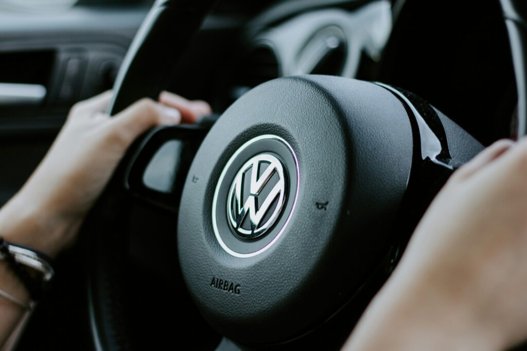 Detalhe do volante de um carro Volkswagen, mostrando o emblema e a inscrição 'Airbag', com as mãos do motorista segurando o volante.