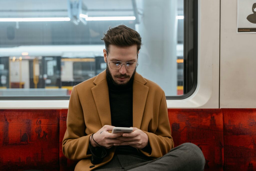 Homem com óculos e casaco de viagem marrom olhando o celular enquanto está sentado em um banco de tecido vermelho dentro de um trem, simbolizando a necessidade de conexão móvel em viagem usando o chip brasileiro no exterior.