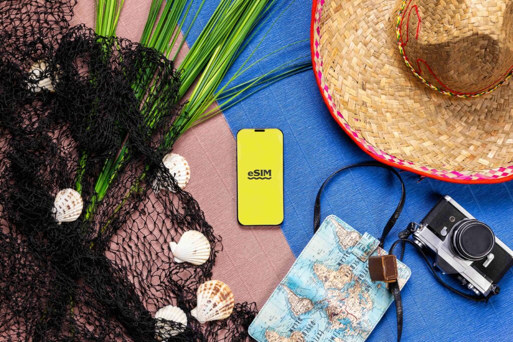 Visão aérea de itens de viagem sobre superfícies coloridas. Os objetos incluem um smartphone com a logo "eSIM" em tela amarela, uma câmera fotográfica vintage, um chapéu de palha, uma rede de pesca preta com conchas, um porta-passaporte com estampa de mapa-múndi e folhas de palmeira.
