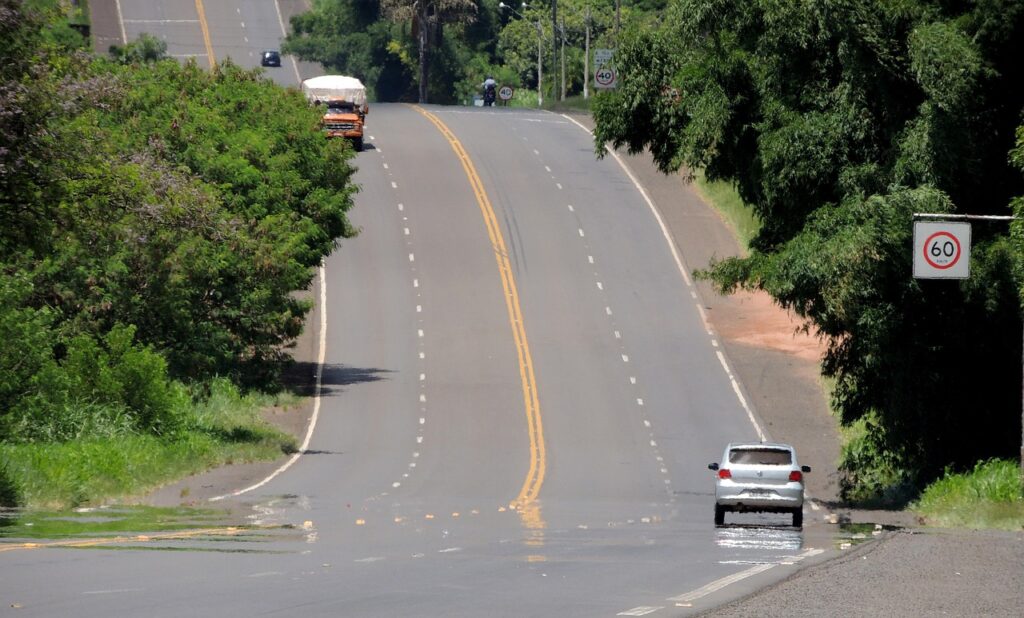 Estrada brasileira em descida e subida com asfalto molhado, cercada por vegetação e com carros em movimento.