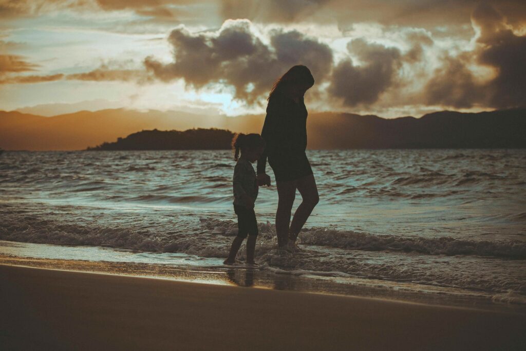 Silhueta de uma mãe e uma criança caminhando juntas na beira do mar durante o pôr do sol em Balneário Camboriú, com reflexos alaranjados na água e o horizonte montanhoso.