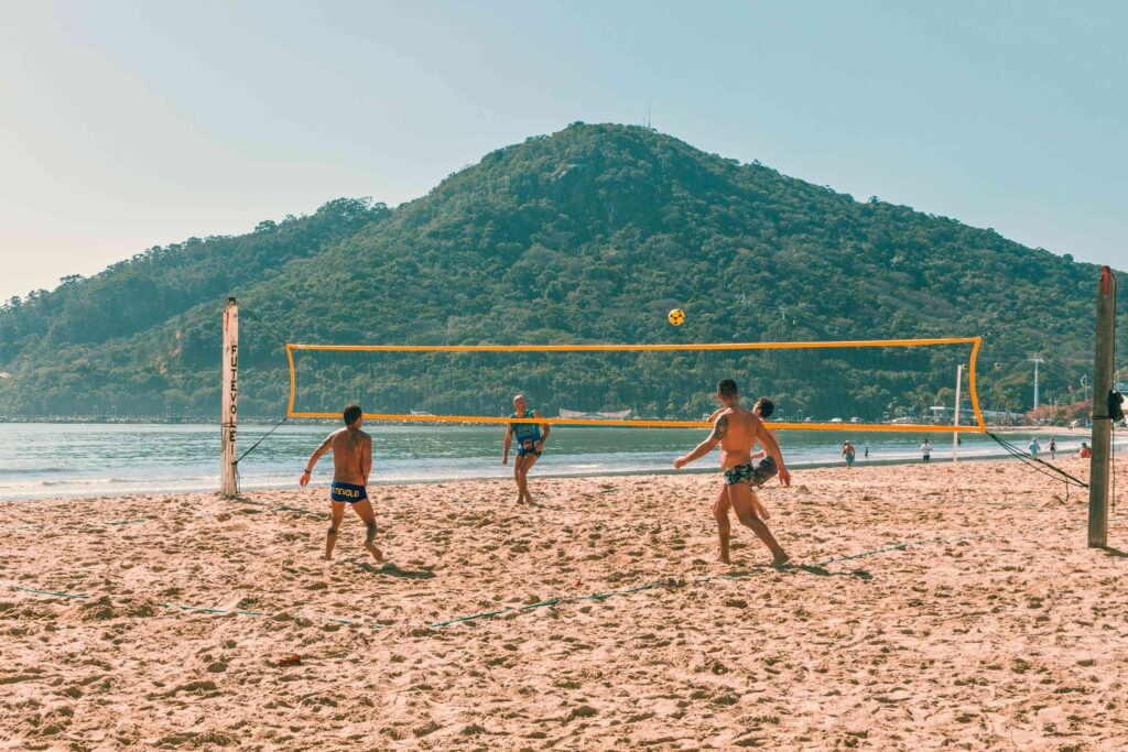 Homens jogando futevôlei na Praia Central de Balneário Camboriú sob sol forte, com mar calmo e o Morro do Careca coberto por vegetação ao fundo.