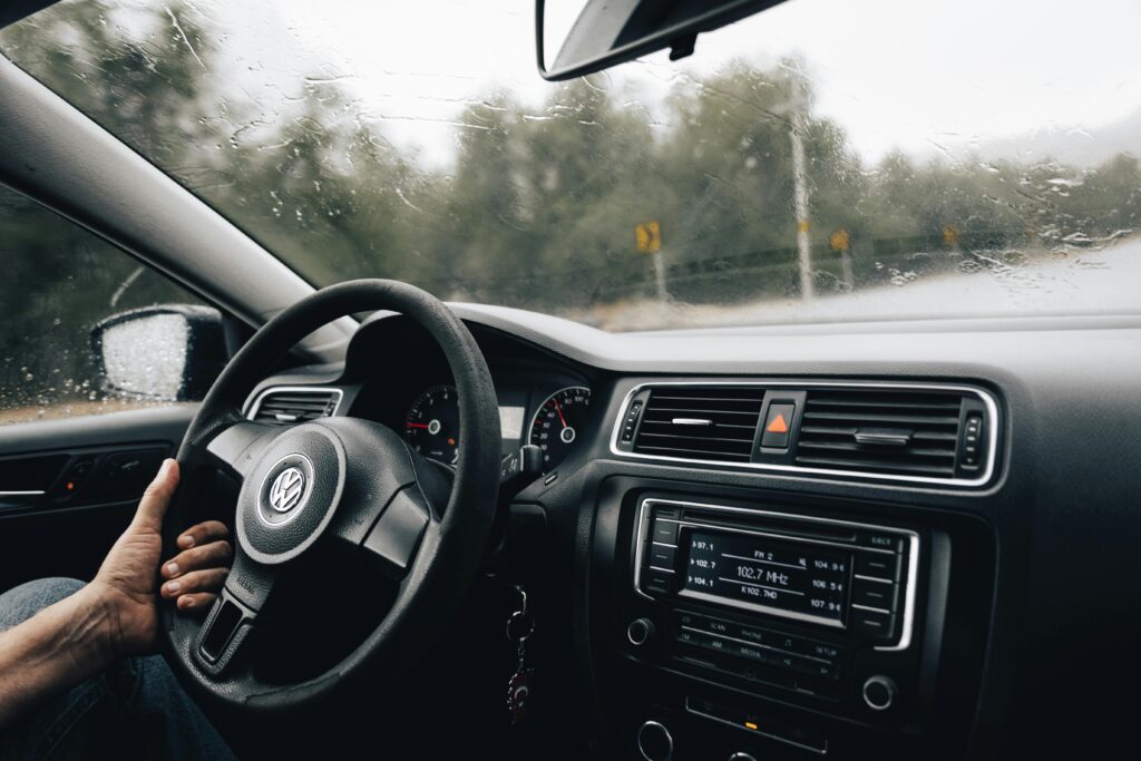 Homem dirigindo um carro em um dia chuvoso, com as mãos no volante. O foco está no interior do carro, mostrando o painel, rádio e parte do para-brisa com gotas de chuva.