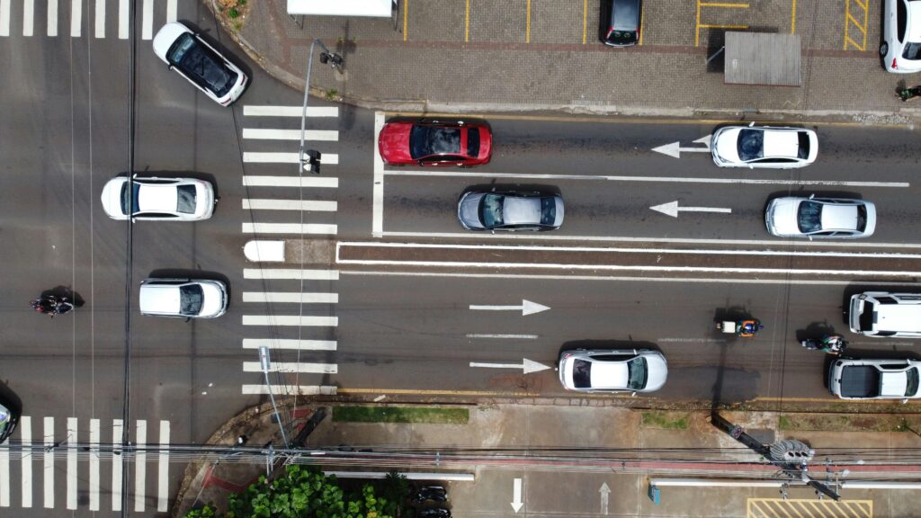 Vista aérea de carros em uma rua movimentada de cidade, mostrando faixas de pedestres e sinalização de trânsito.