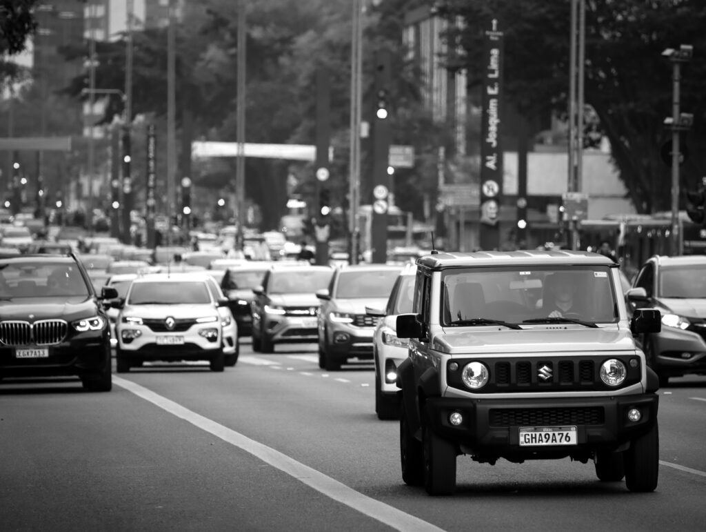 Foto em preto e branco de um congestionamento em uma avenida movimentada, com diversos carros alinhados em quatro faixas. Um Suzuki Jimny em primeiro plano se destaca na rua, com prédios e árvores em segundo plano.