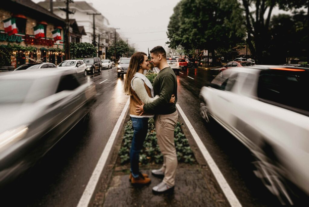 Casal de mãos dadas, sorrindo e abraçado, parado em canteiro central de rua (Avenida Borges de Medeiros) com asfalto molhado e carros em movimento borrado (efeito de velocidade), ressaltando o clima romântico e urbano de Gramado.