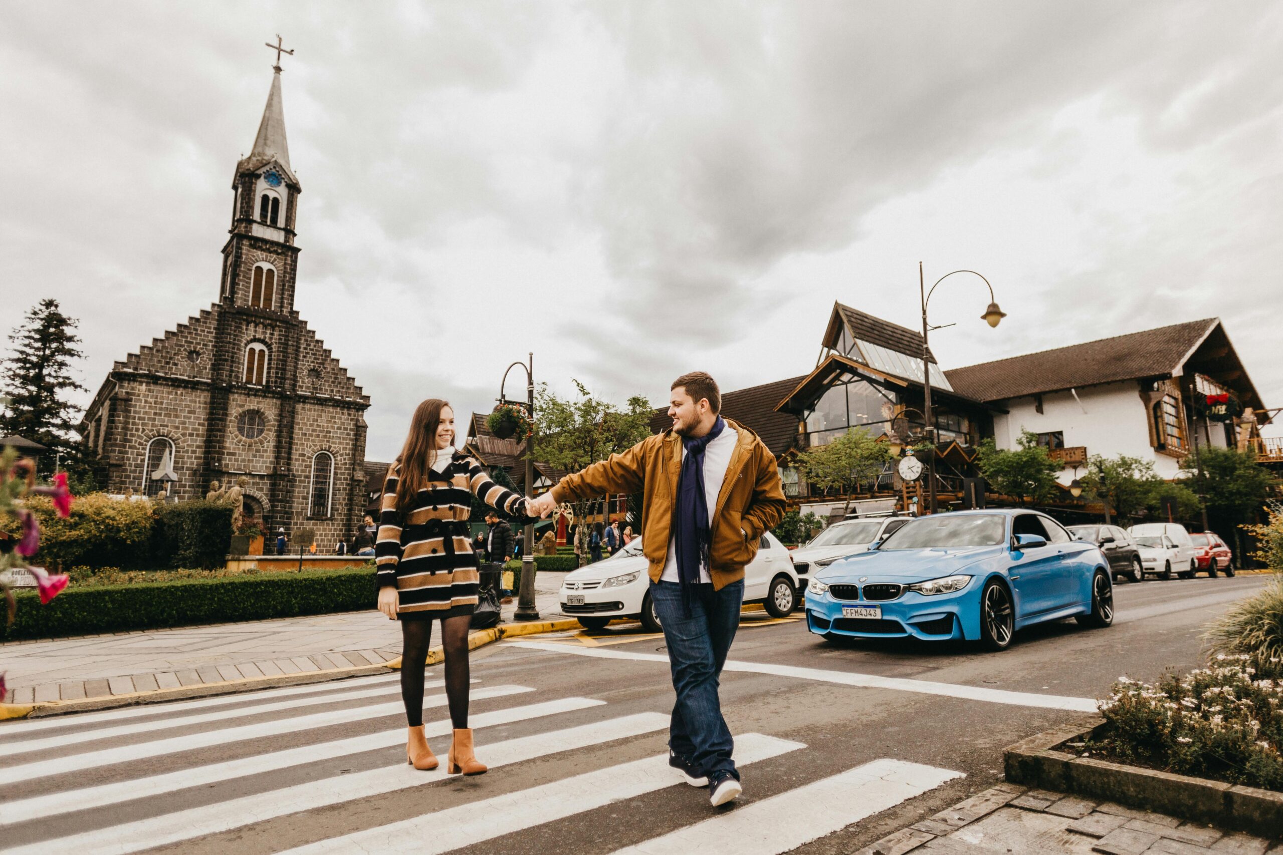 Casal de turistas (homem e mulher) de mãos dadas atravessando faixa de pedestres em frente à Igreja Matriz São Pedro Apóstolo em Gramado, com a fachada de pedra em destaque e céu nublado, realçando o clima europeu da Serra Gaúcha. Foto: Jonathan Borba/Pexels