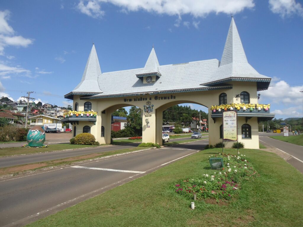 Pórtico de Gramado RS na entrada pela RS-115 (Via Taquara) com estilo normando e torres pontiagudas, cartão-postal da cidade, em dia de sol com céu azul e vegetação verde nas laterais da rodovia.