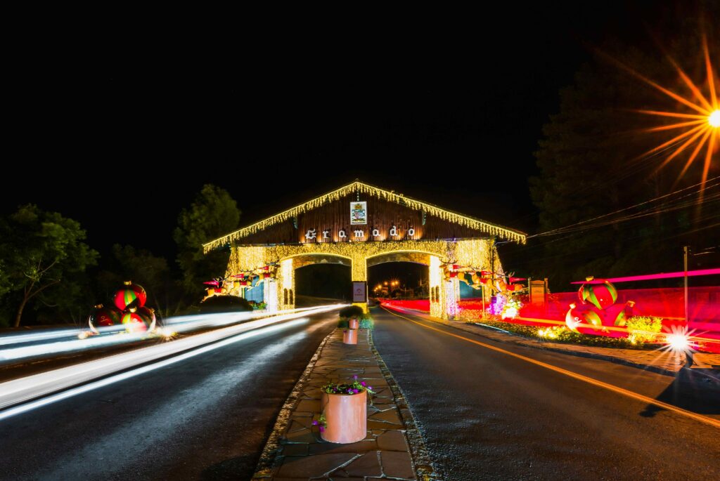 Pórtico de Gramado (Via Nova Petrópolis) à noite, totalmente iluminado com luzes de Natal amarelas; em primeiro plano, rastros de luz branca e vermelha de veículos em movimento na rodovia, indicando o trânsito da entrada da cidade.