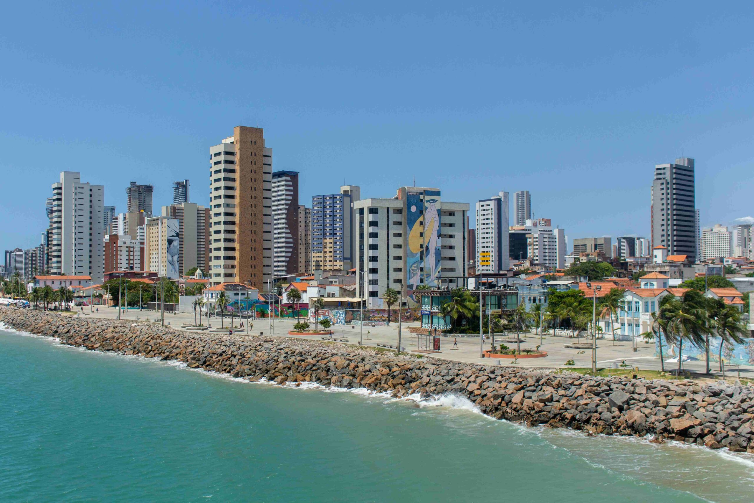 Skyline e orla da Praia de Iracema, Fortaleza, Ceará, mostrando prédios altos, o quebra-mar de pedras e a cor esmeralda do mar em um dia ensolarado.