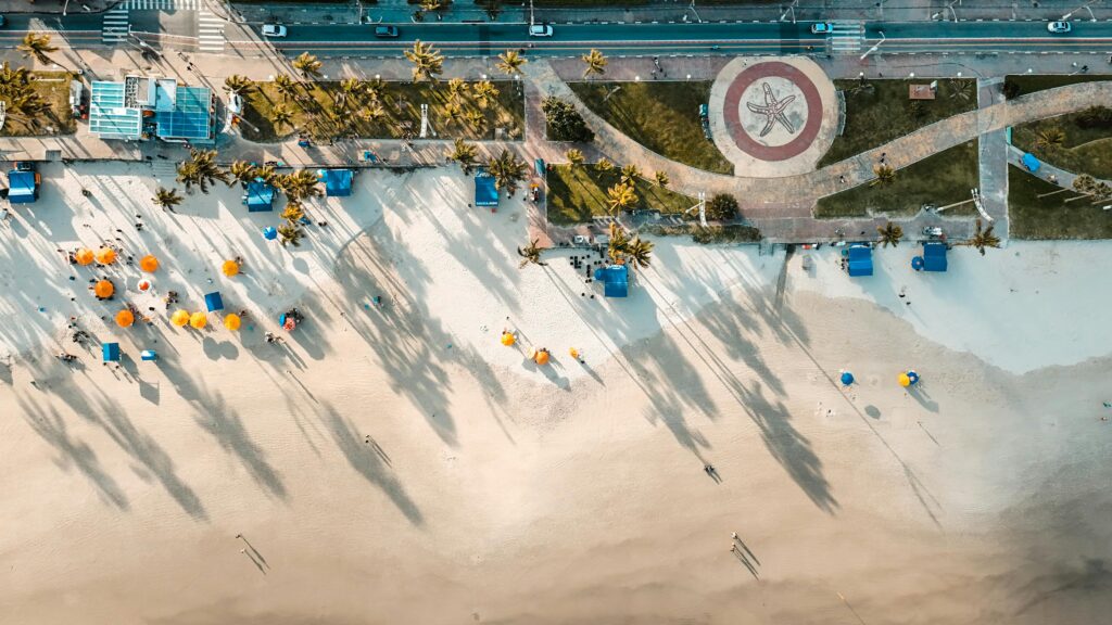 Drone capturando a Praia de Iracema em Fortaleza, Ceará, com palmeiras, guarda-sóis coloridos e a avenida beira-mar, destacando o contraste de luz e sombra na areia.