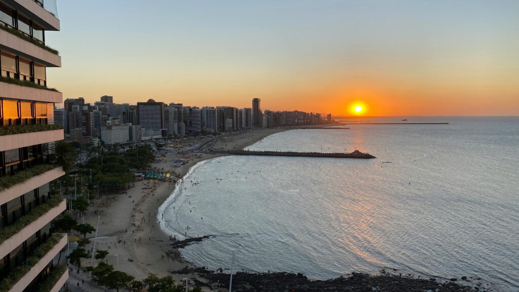 Pôr do sol panorâmico em Fortaleza, Ceará, mostrando a Praia de Meireles cheia de pessoas, o píer de Iracema e os prédios da orla urbana ao entardecer.