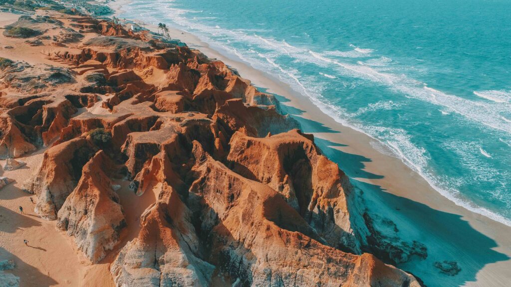 Vista aérea das falésias de Morro Branco em tons de laranja e vermelho, contrastando com a areia clara da praia e o mar azul-turquesa do Ceará.