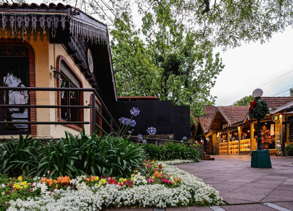 Canteiro de flores coloridas em primeiro plano com foco em uma casa de arquitetura típica europeia em Gramado, com telhado escuro e detalhes em madeira (estilo enxaimel), em uma rua charmosa da Serra Gaúcha.