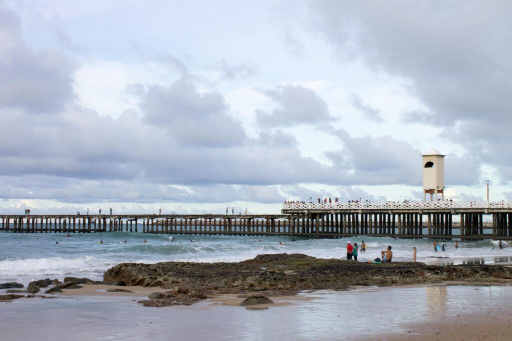 Píer ou espigão de Iracema em Fortaleza, Ceará, sobre o mar com pessoas caminhando, uma torre de observação na ponta e banhistas na praia rochosa em primeiro plano.
