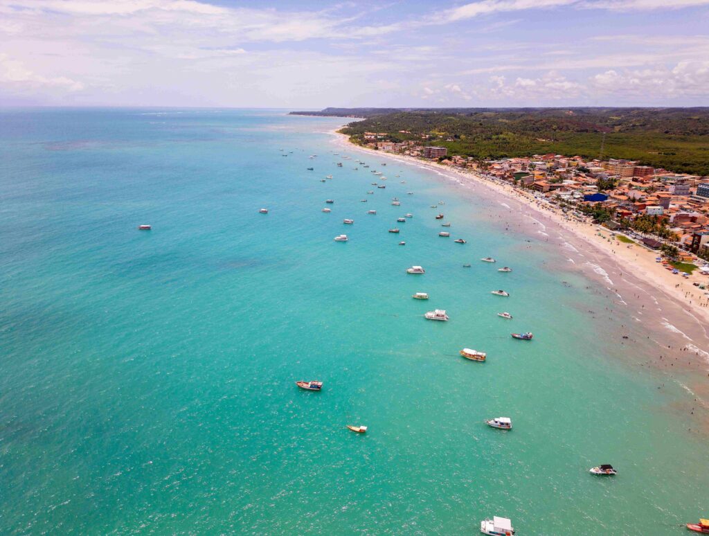 Foto aérea da orla de Maragogi, Alagoas, mostrando o mar azul-claro com grande quantidade de barcos e catamarãs próximos à praia, indicando o embarque para as piscinas naturais.